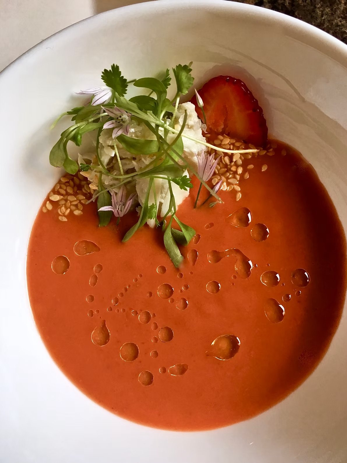 A bowl of soup garnished with microgreens, a dollop of cream, sesame seeds, and a sliced strawberry.