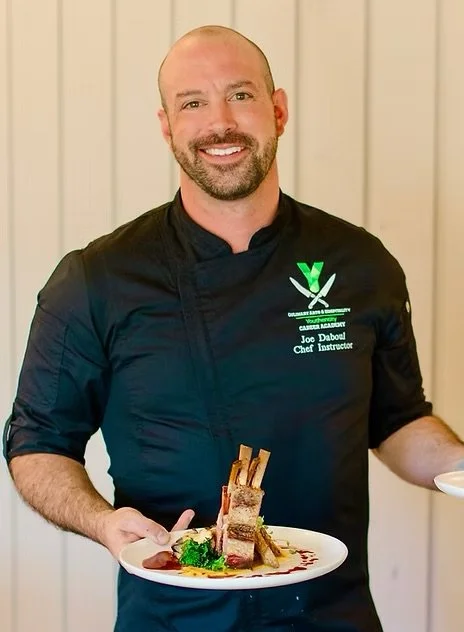 A smiling man in a black chef's coat with green logo and name tag holding a plate of sliced meat with sauce and vegetables, standing in front of a wooden wall.