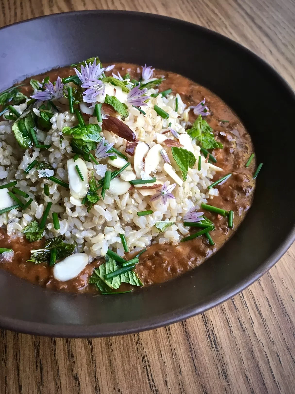 A bowl of rice with herbs and edible flowers on top, sitting on a wooden surface.