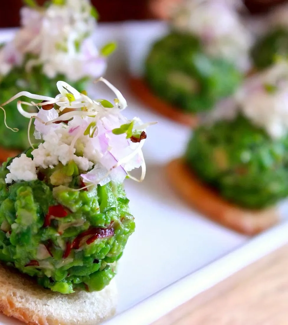 Close-up of a gourmet appetizer featuring english peas topped with microgreens and edible flowers, served on a bread base with other similar topped bread pieces in the background.