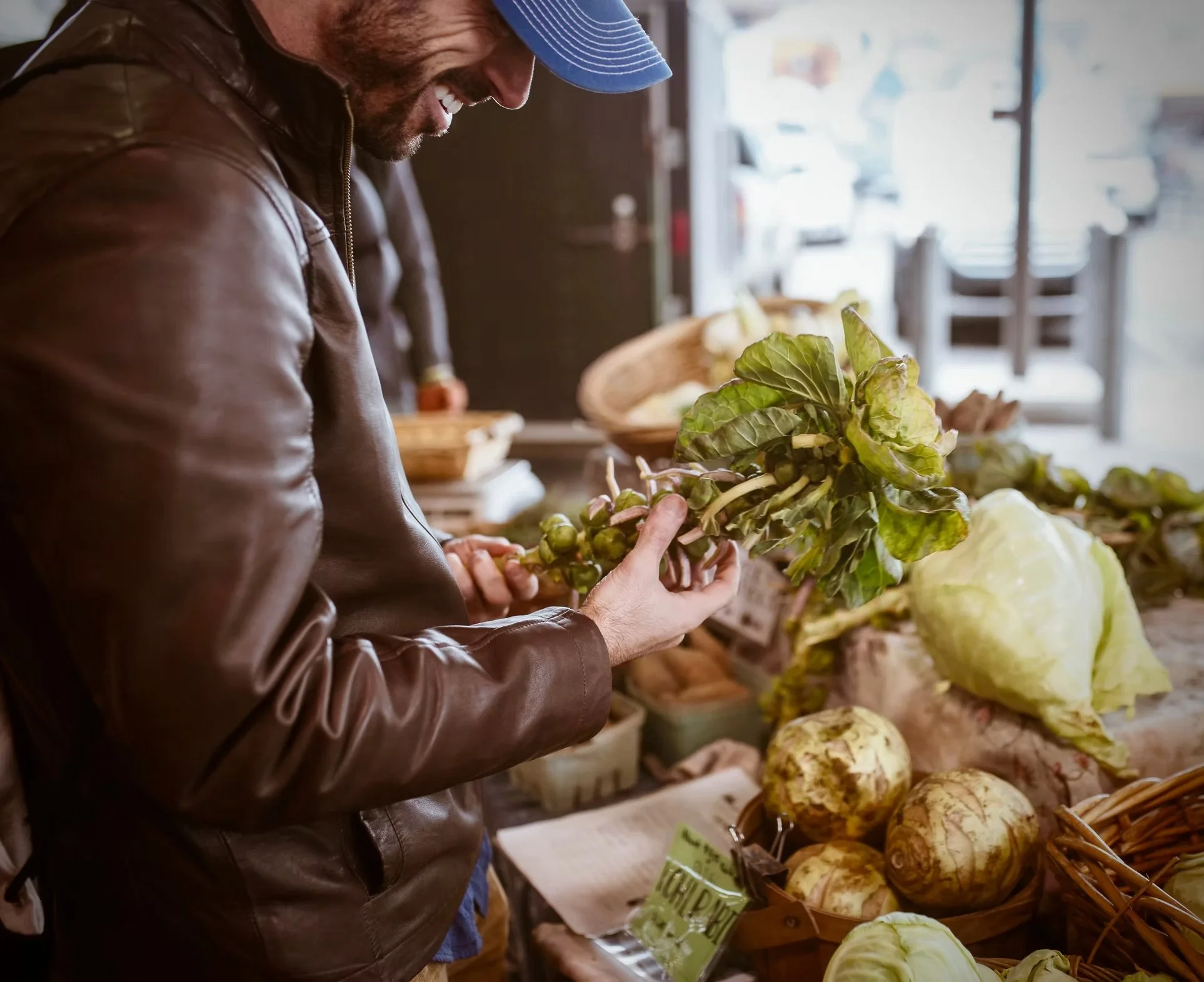 A man wearing a brown leather jacket and a blue striped cap shopping for fresh vegetables at an outdoor market, holding a bunch of leafy greens.