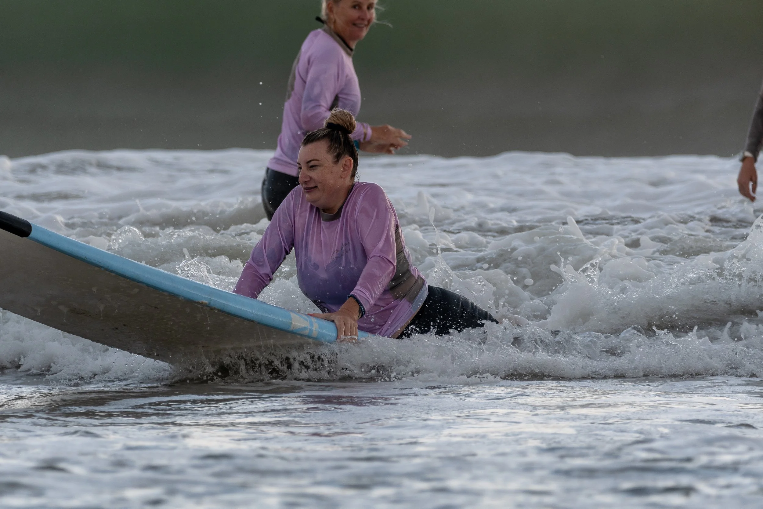 Two women in purple shirts are on a surfboard in the water, with one sitting and the other standing behind her, both smiling and enjoying the moment.