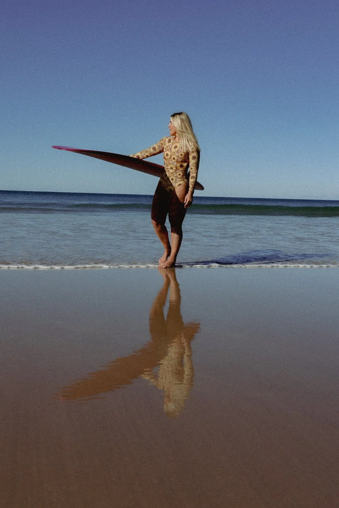 A woman with long blonde hair standing on a beach holding a surfboard, wearing a floral top and black wetsuit pants, with her reflection visible on the wet sand.
