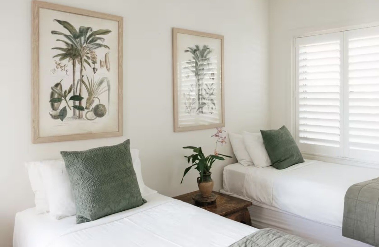 Two beds with white bedding and green pillows in a bright room with framed botanical prints and a potted plant on a wooden table between the beds.