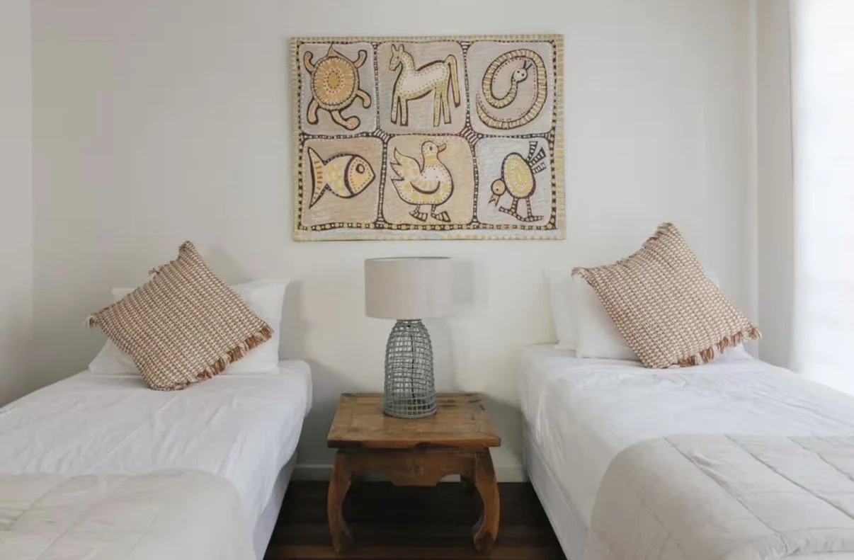 A bedroom with two single beds with white bedding and checkered pillows, a wooden nightstand with a lamp between them, and a colorful animal-themed artwork on the wall.