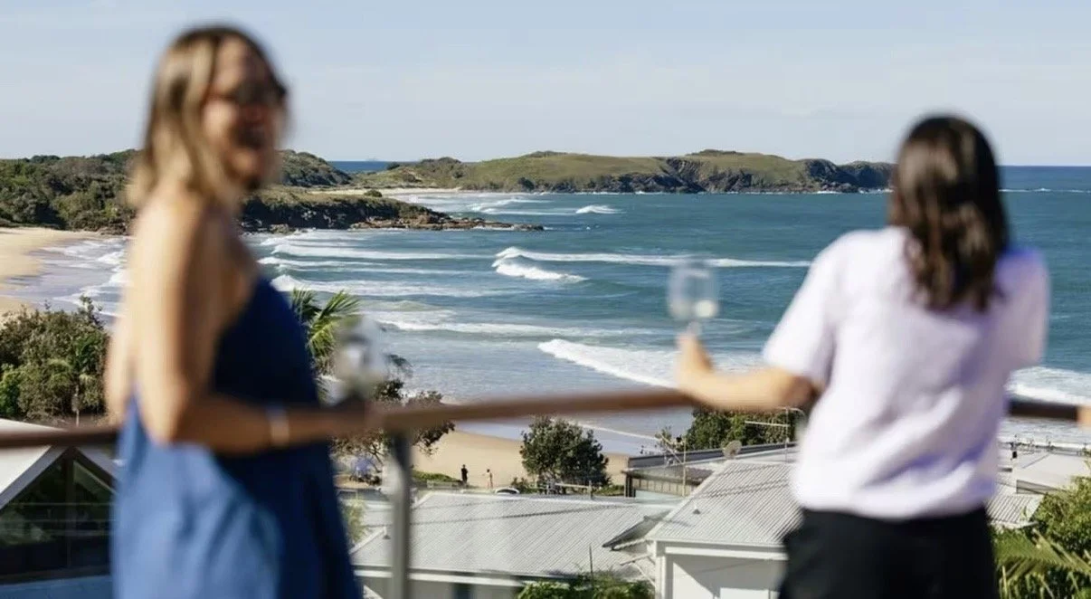 Two women holding wine glasses on a balcony overlooking a beach and ocean with waves and green islands in the distance.