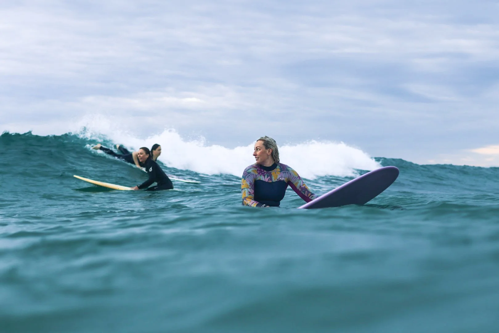 A woman in a wetsuit with a colorful pattern on the sleeves holding a paddleboard in the ocean while two other surfers in black wetsuits paddle on surfboards further behind in the water, under a cloudy sky.