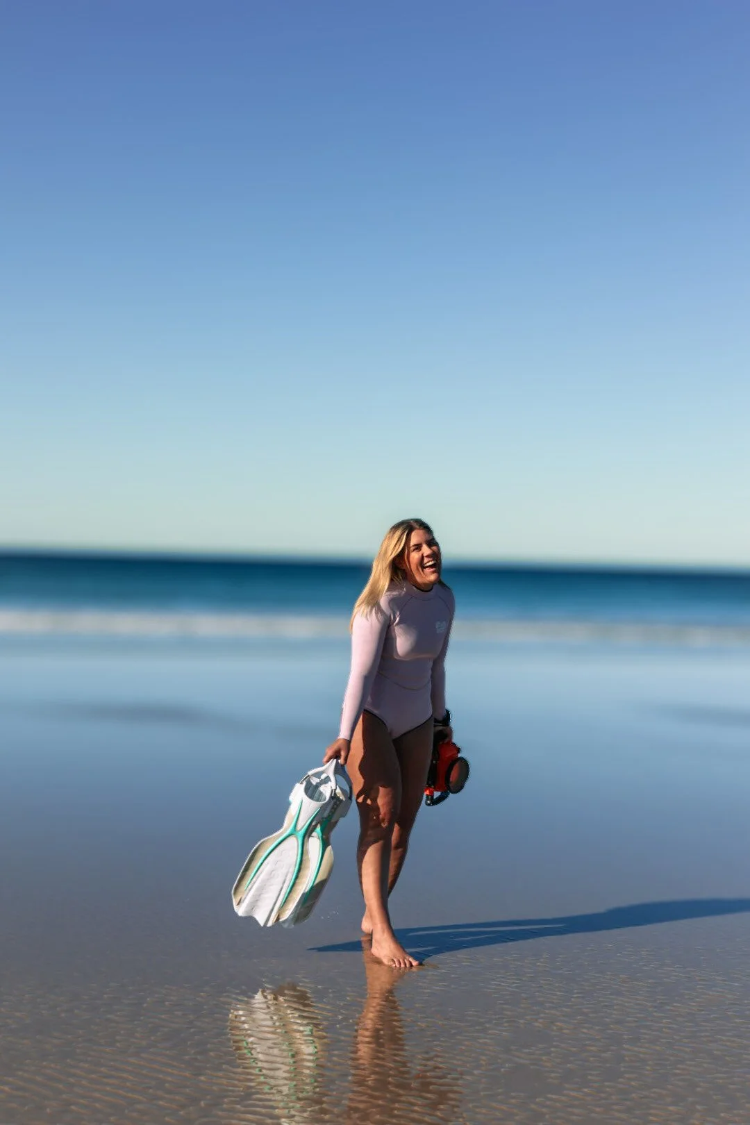 Woman in wetsuit smiling at the beach, holding fins in one hand and snorkeling gear in the other, standing barefoot on wet sand with ocean and clear sky in the background.