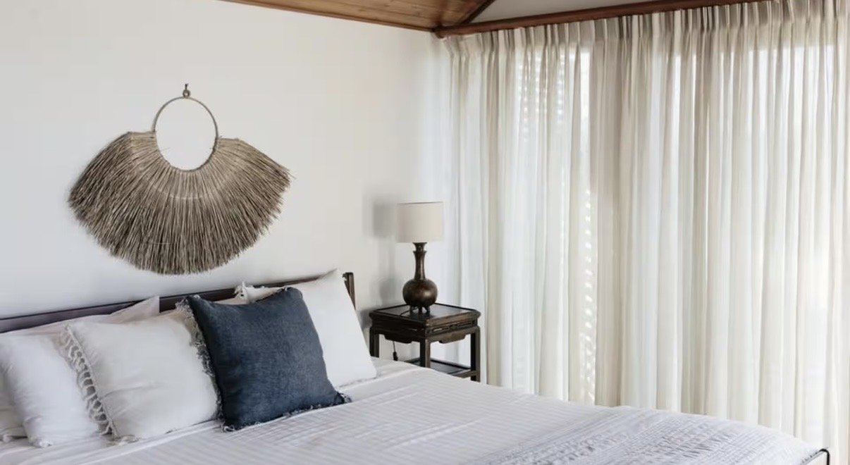 A neatly made bed with white linens and pillows, a dark wooden nightstand with a lamp, and a decorative wall hanging made of woven straw above the bed. Cream curtains cover large windows in a cozy bedroom with wooden ceiling.