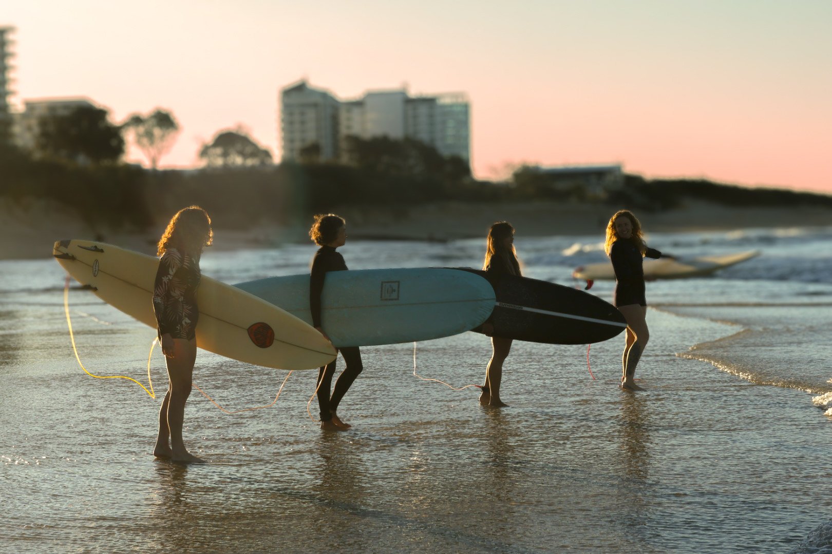 Four women carrying surfboards walk into the ocean during sunset on a beach, with buildings in the background.