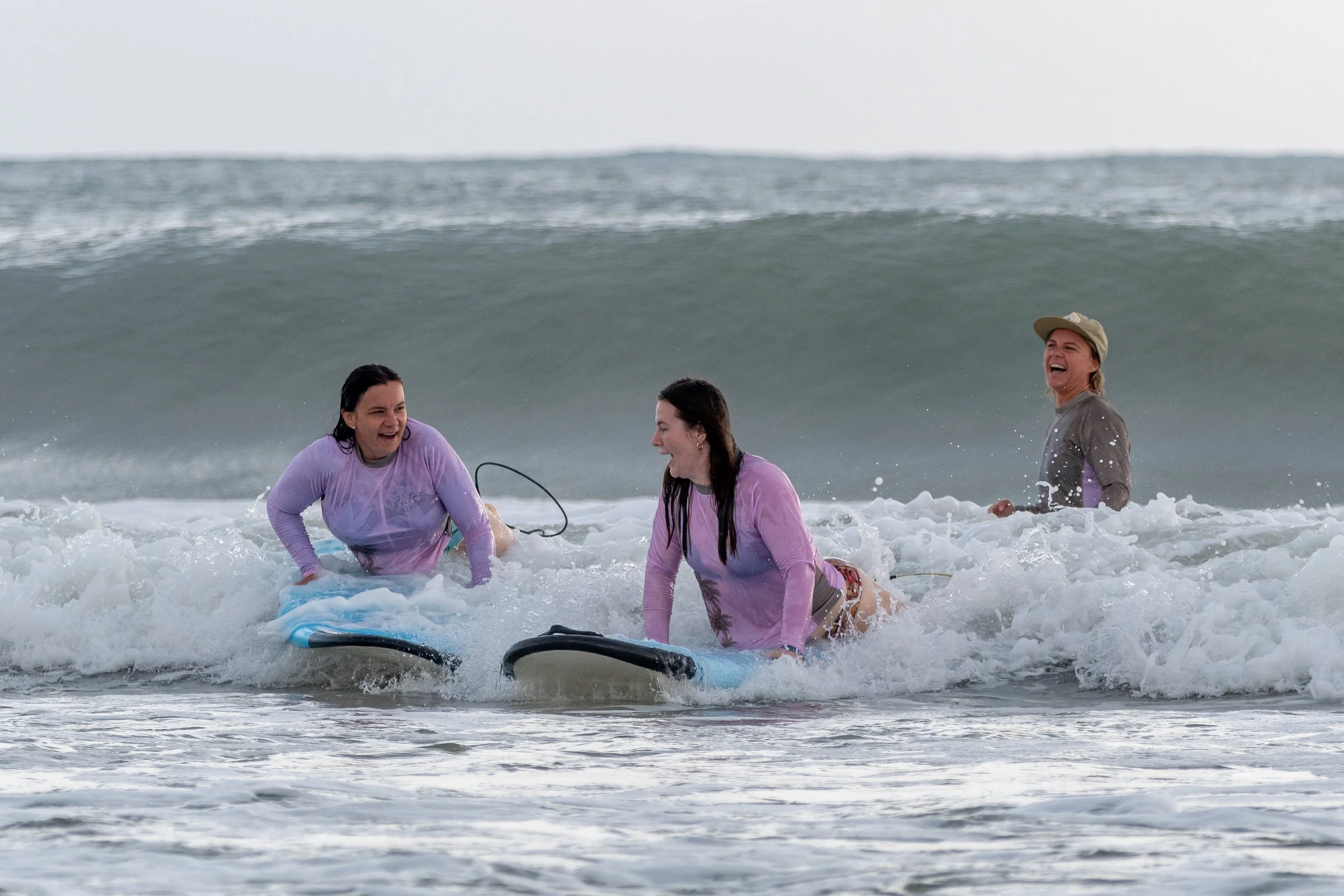 Four women enjoying a day at the beach, playing in the ocean waves with surfboards, smiling and laughing.