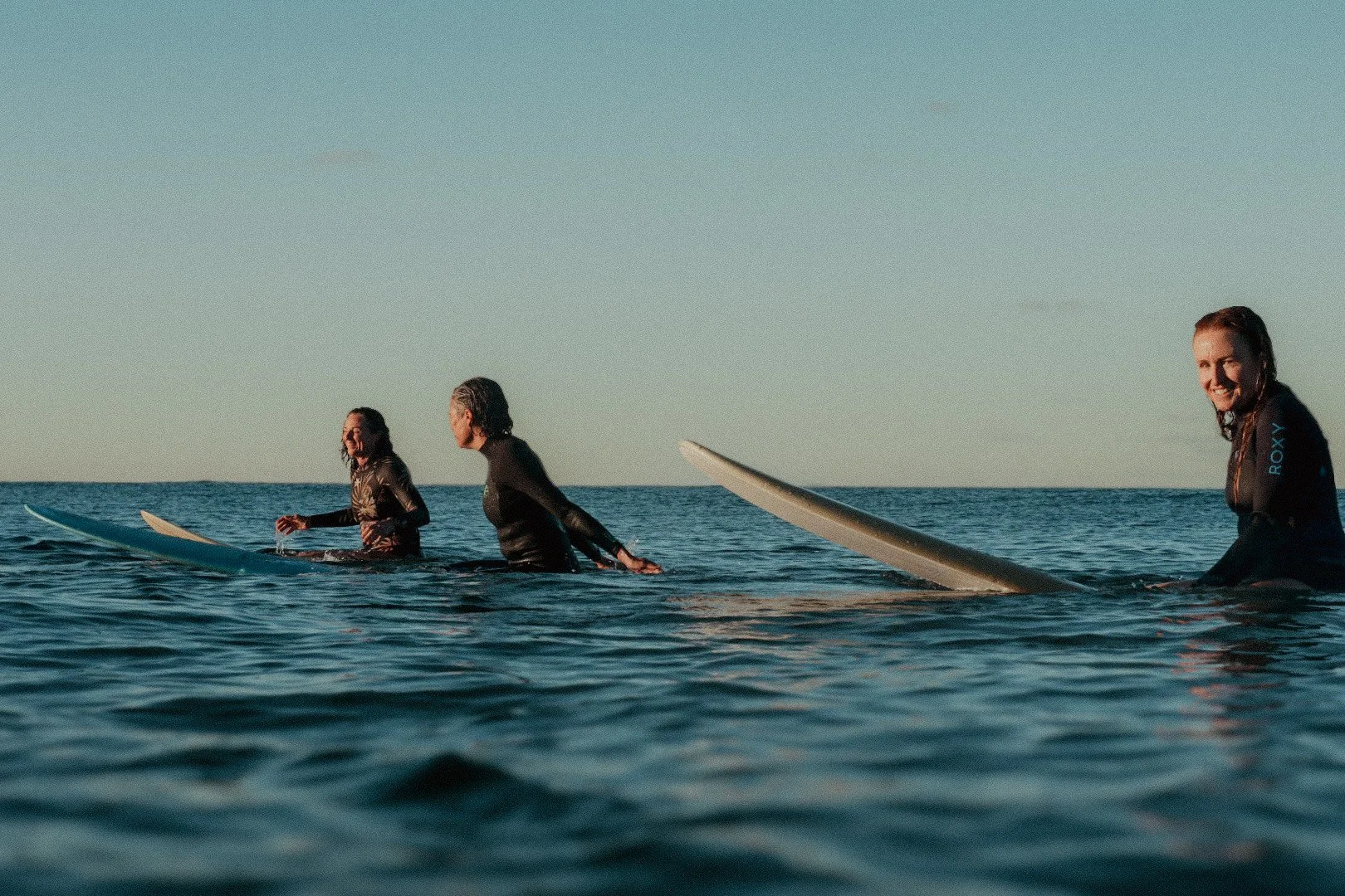 Four women in wetsuits sitting on surfboards in the ocean, smiling and enjoying a sunny day.