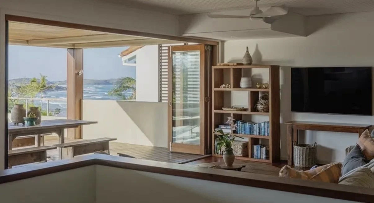 Living room with sliding glass doors open to a balcony overlooking the ocean, with palm trees and a blue sky, wooden furniture, a TV, and decor.