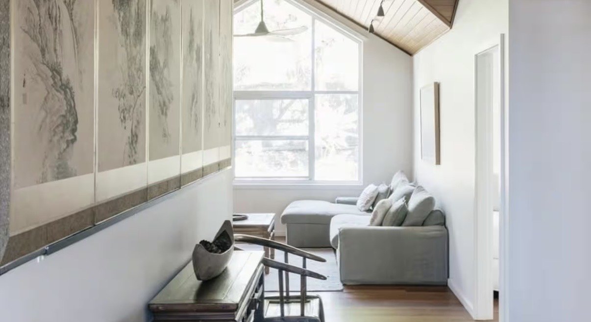 Bright living room with a large window, a white sectional sofa with pillows, wood-paneled ceiling, artwork on the walls, and a wooden bench with a decorative bowl.