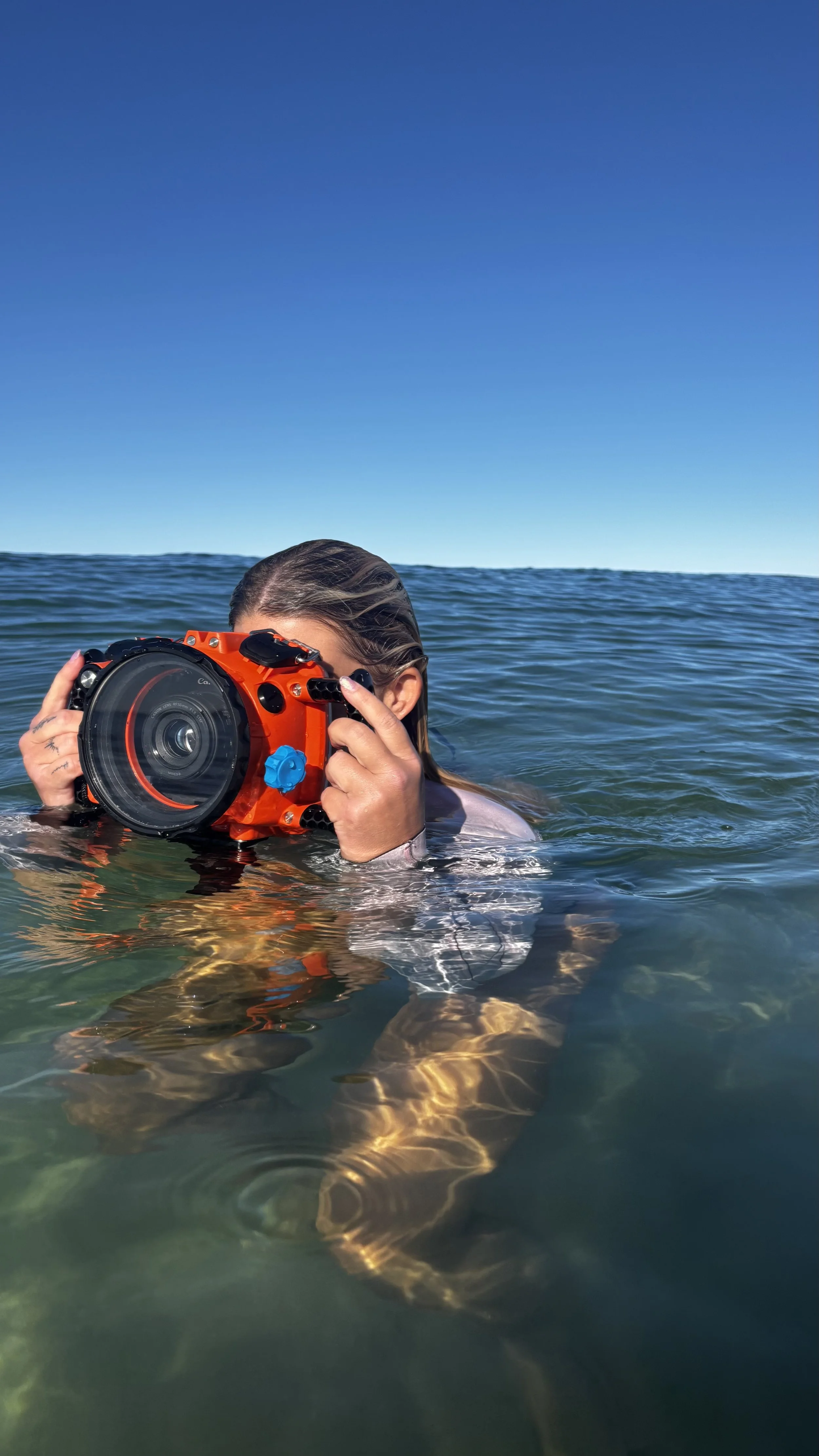 A person with wet hair in the ocean holding a large orange underwater camera.