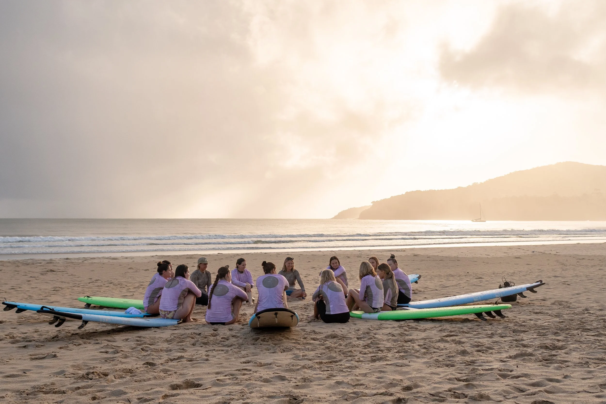 A group of people gathering on a sandy beach, with surfboards lying on the sand nearby. The sky is cloudy with sunlight breaking through, and the ocean waves are visible in the background. Mountainous terrain can be seen in the distance.