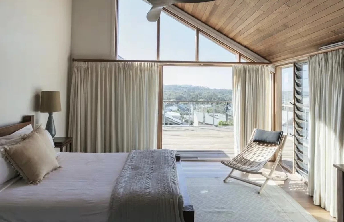 Bedroom with a bed on the left side, beige pillows, beige curtains covering a large window, a wooden ceiling, a chair near the window, and a view of rooftops and trees outside.