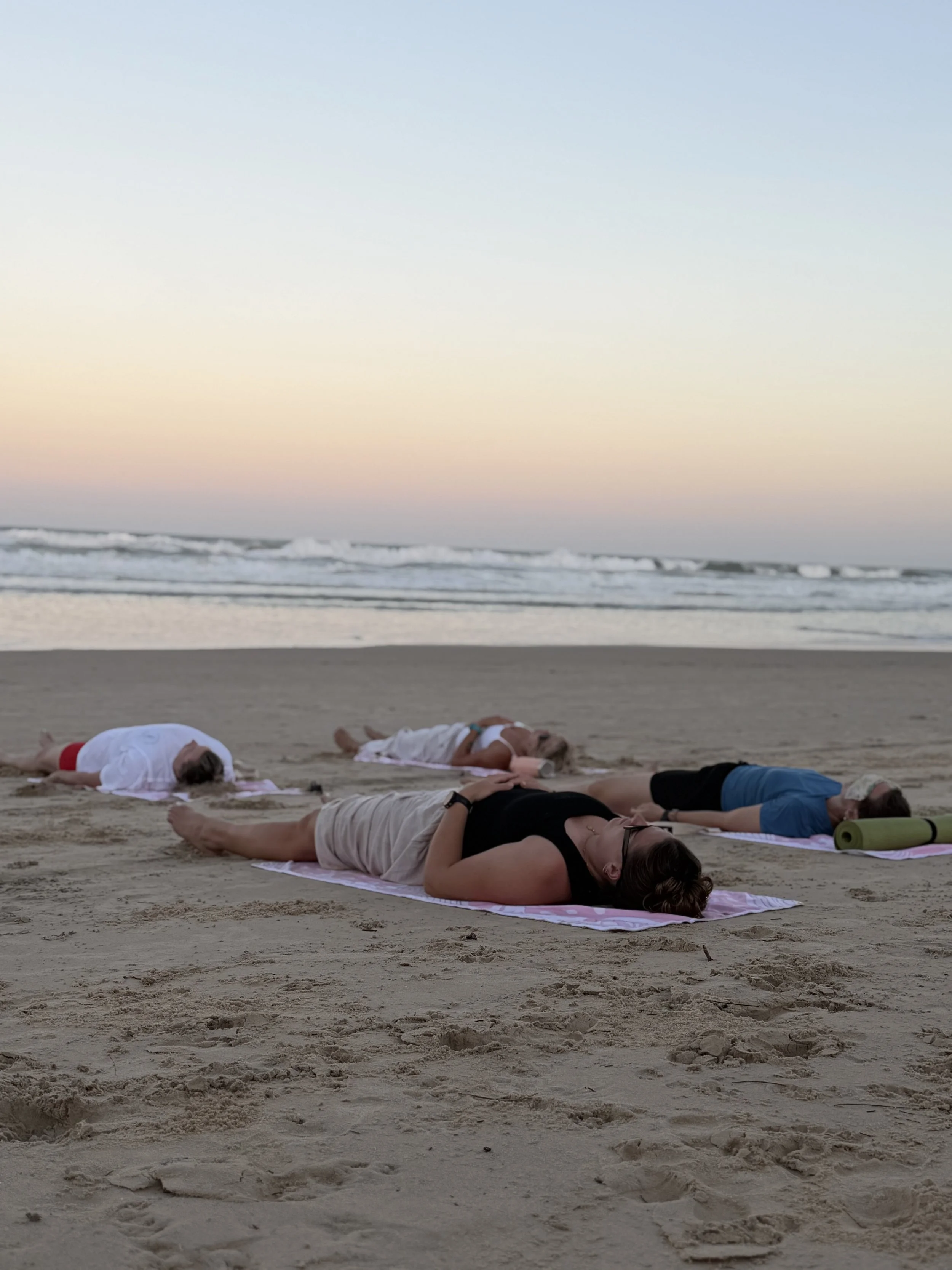 People practicing yoga on a sandy beach during sunset, lying on their stomachs with arms extended forward and faces resting on the ground.