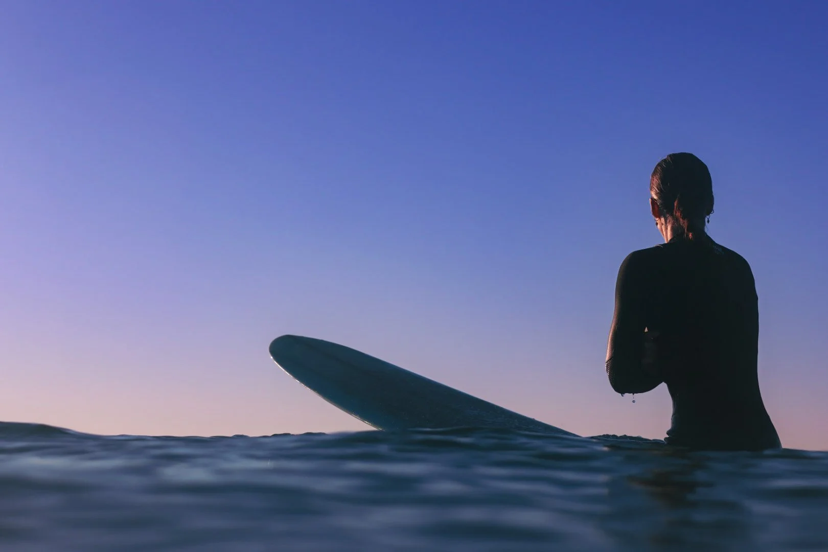 A person sitting on a surfboard in the water during sunset, facing away from the camera with a clear sky.