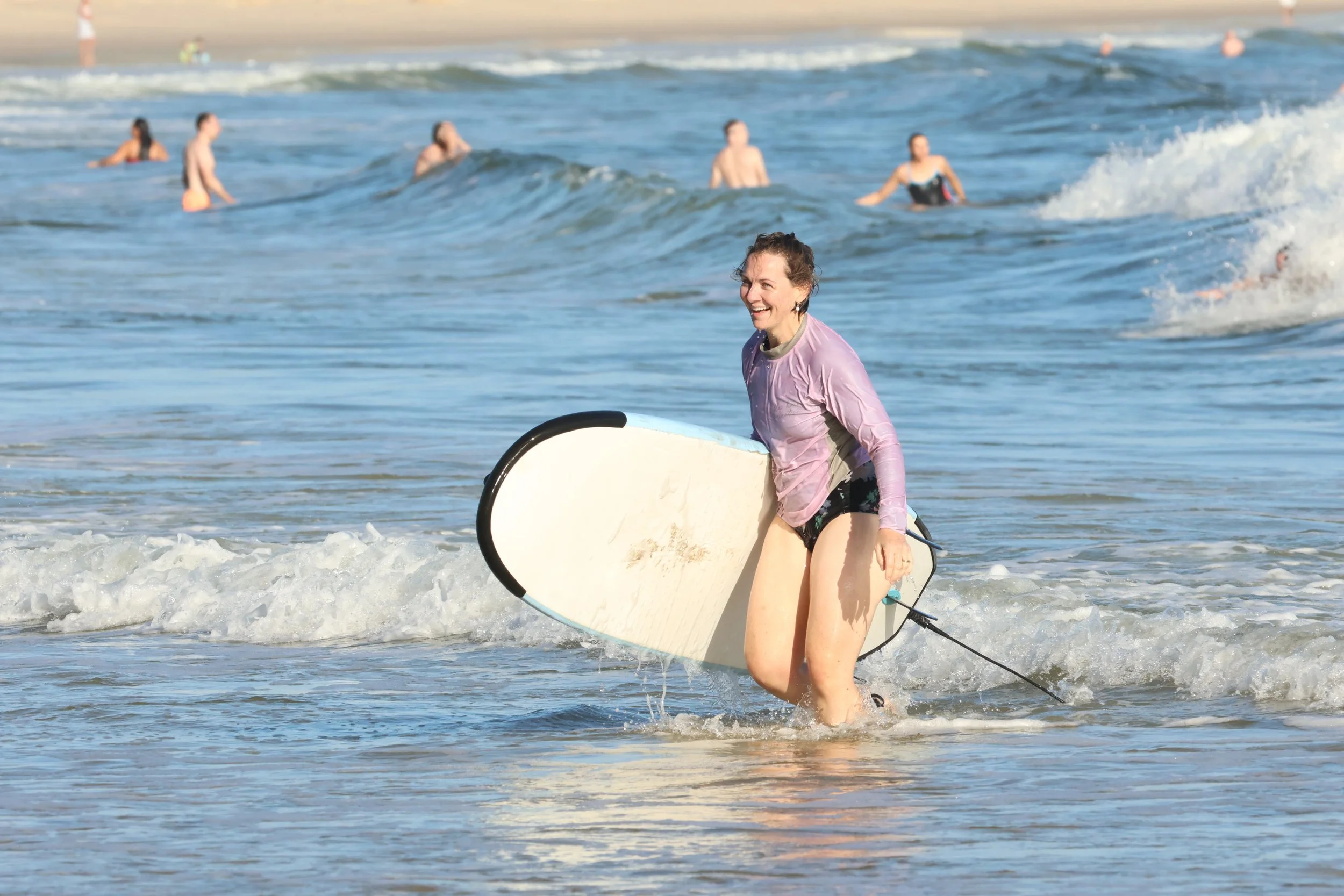 A woman with a surfboard in the ocean, smiling, while other surfers are in the water behind her, with a sandy beach in the background.