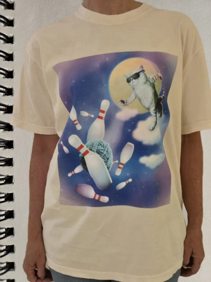 A person wearing a cream-colored T-shirt with a graphic of a cat wearing sunglasses, bowling, among clouds and a large yellow moon in space.