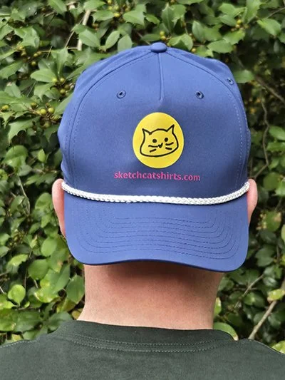 Blue baseball cap with a yellow circle featuring a cat face, and pink text that reads 'sketchcatshirts.com' in front of green foliage.