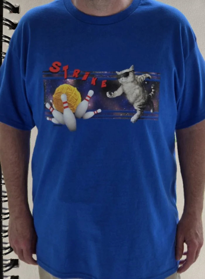 Blue T-shirt with a graphic of a yarn ball, pins and a cat in space bowling, with the word 'Strike' in red letters.