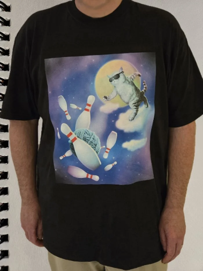 T-shirt with a graphic of a cat wearing sunglasses bowling and floating in space with a knitting ball and bowling pins against a full moon and starry sky background.