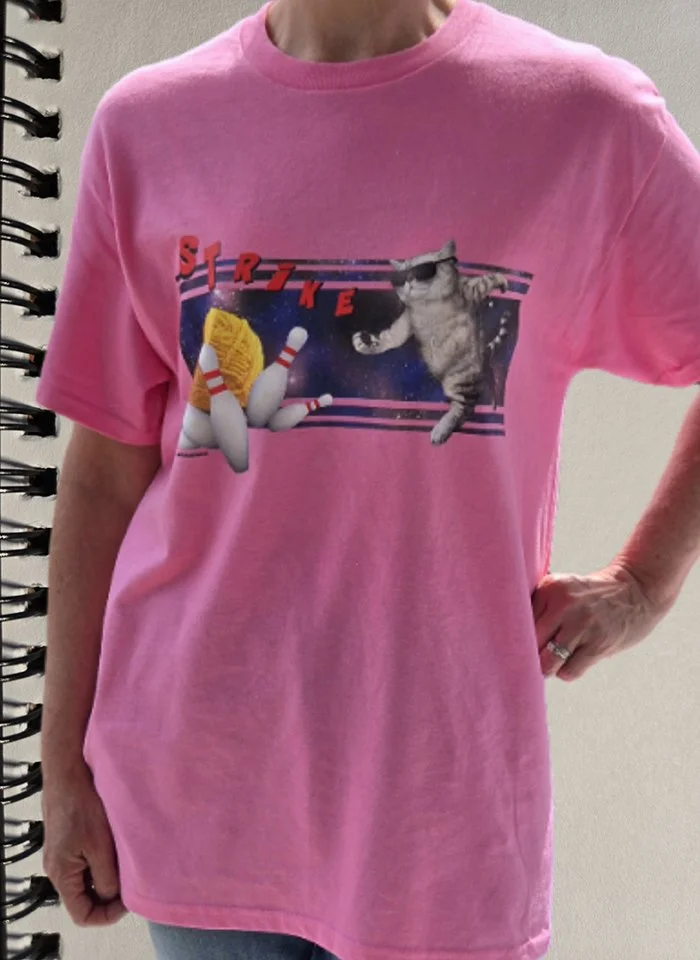 Pink T-shirt with a graphic of a yarn ball, pins and a cat in space bowling, with the word 'Strike' in red letters.