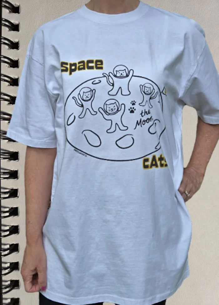 A person is wearing a white T-shirt with a cartoon illustration of cats on the moon, with the text 'space cats' and 'the moon'.