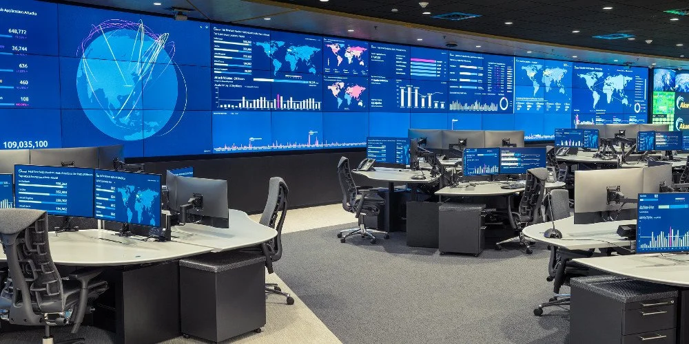 A control room with multiple desks, each having computer monitors displaying charts, graphs, and maps. Large digital screens on the wall show data visualizations, world maps, and analytics in a high-tech environment.