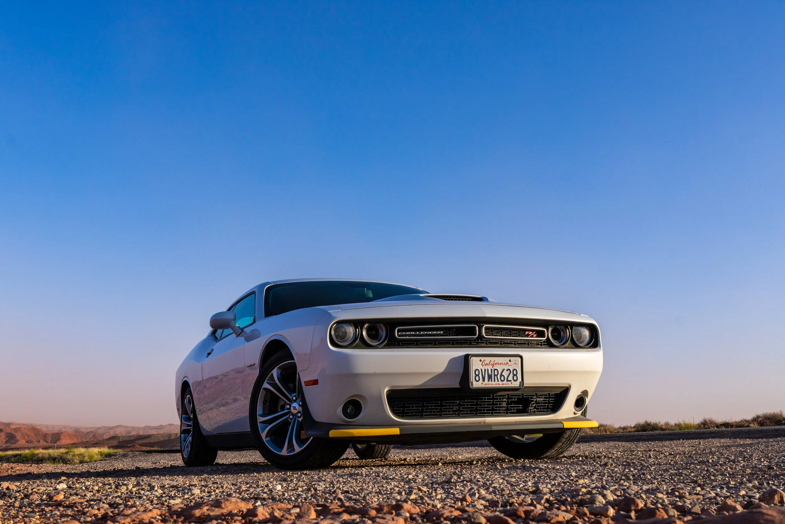 A white Dodge Challenger R/T parked on a gravel road in a desert landscape under a clear blue sky.