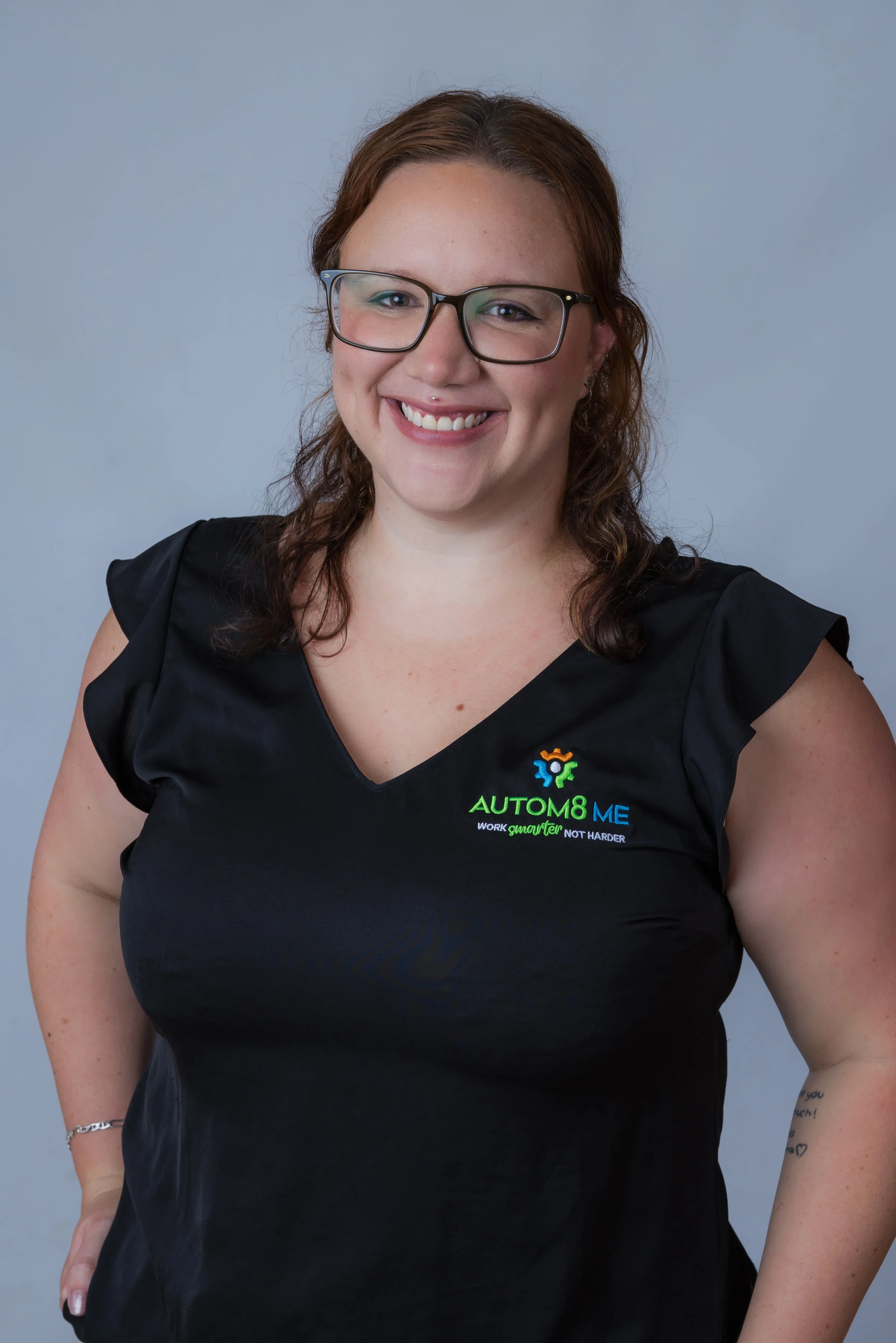 A woman with curly brown hair and glasses, smiling, wearing a black shirt with a logo that reads 'AUTOM8 ME' and the slogan 'Work smarter not harder'.