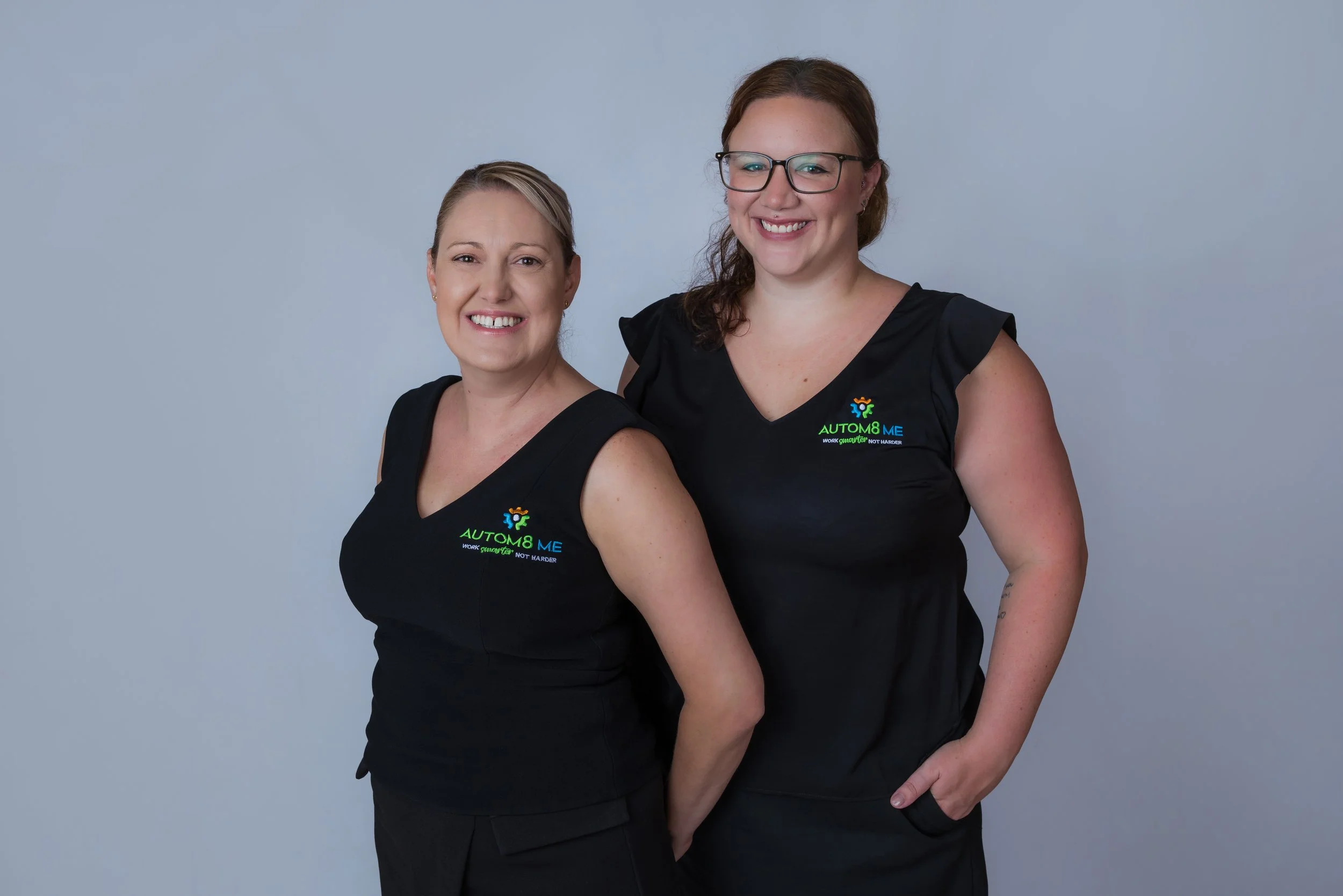 Two smiling women wearing black scrubs with 'Autom8 Me' text standing against a light grey background.