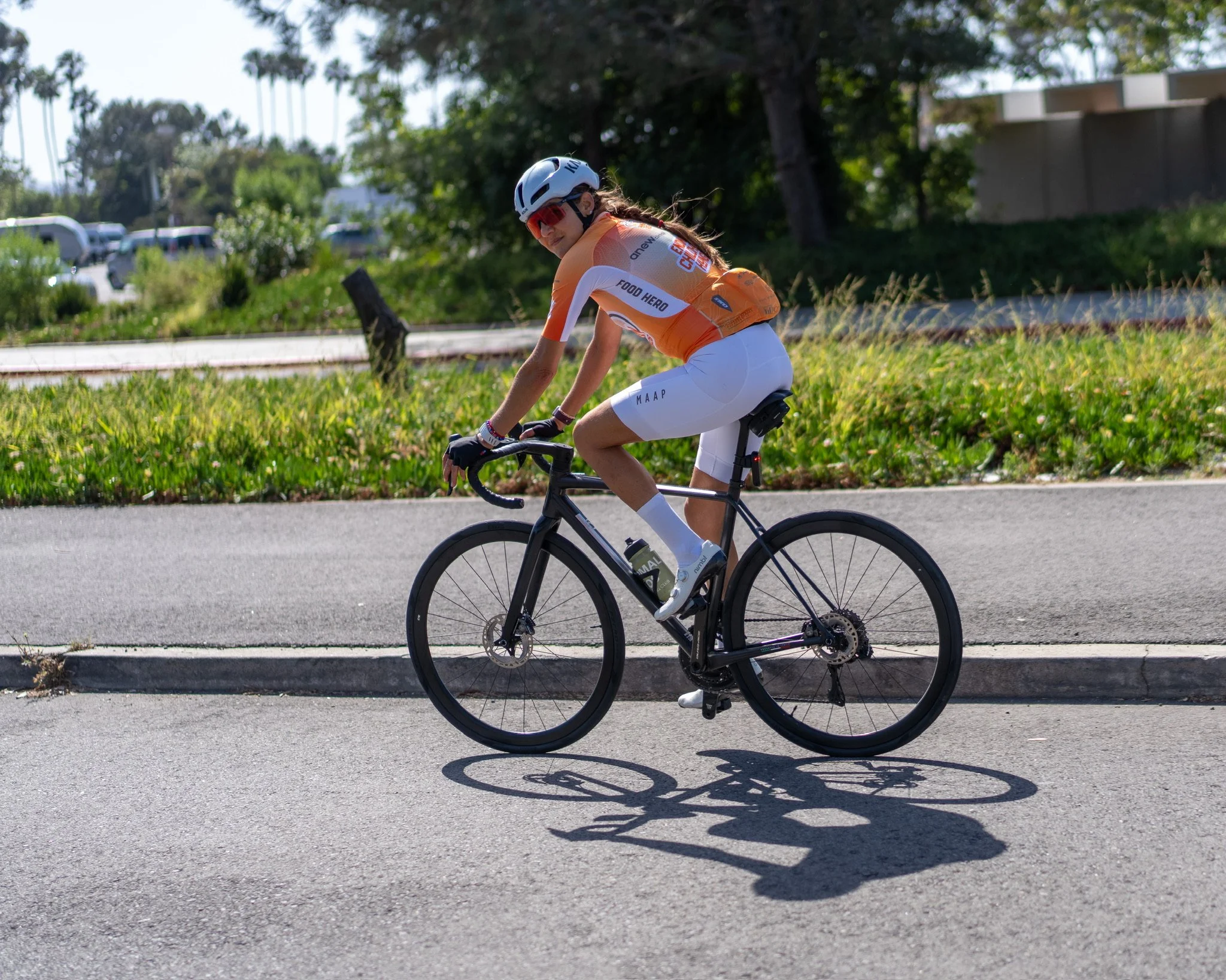 A woman wearing a helmet and cycling gear rides a black road bike on a paved path, with greenery and parked cars in the background.