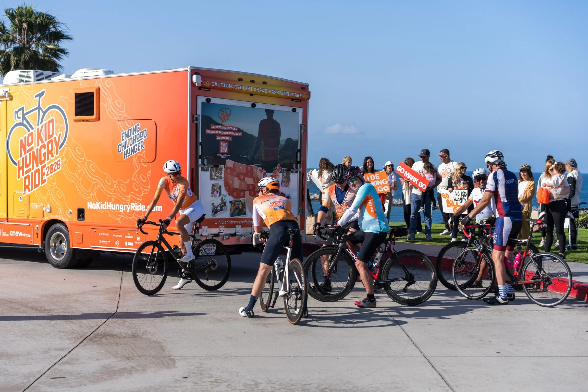 A group of cyclists participating in a charity bike ride event called 'No Kid Hungry Ride,' with a bright orange mobile support vehicle, some carrying signs, and people gathered by the waterfront on a sunny day.