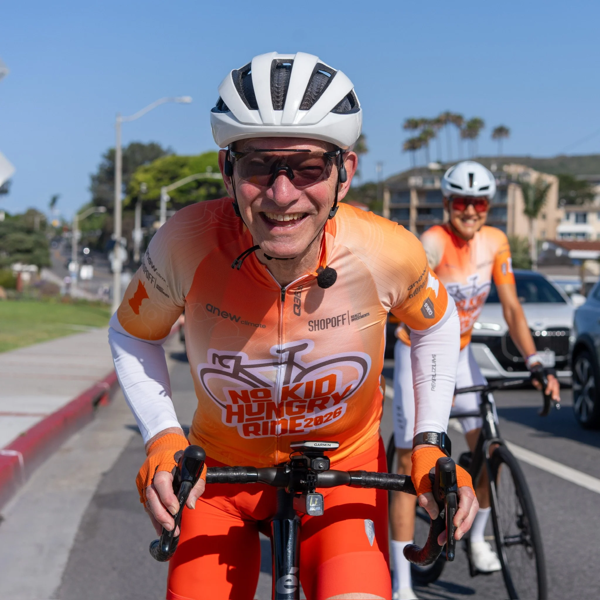 Close-up of a smiling man wearing a white bike helmet and orange cycling jersey, riding a bicycle outdoors with other cyclists visible in the background under a clear blue sky.