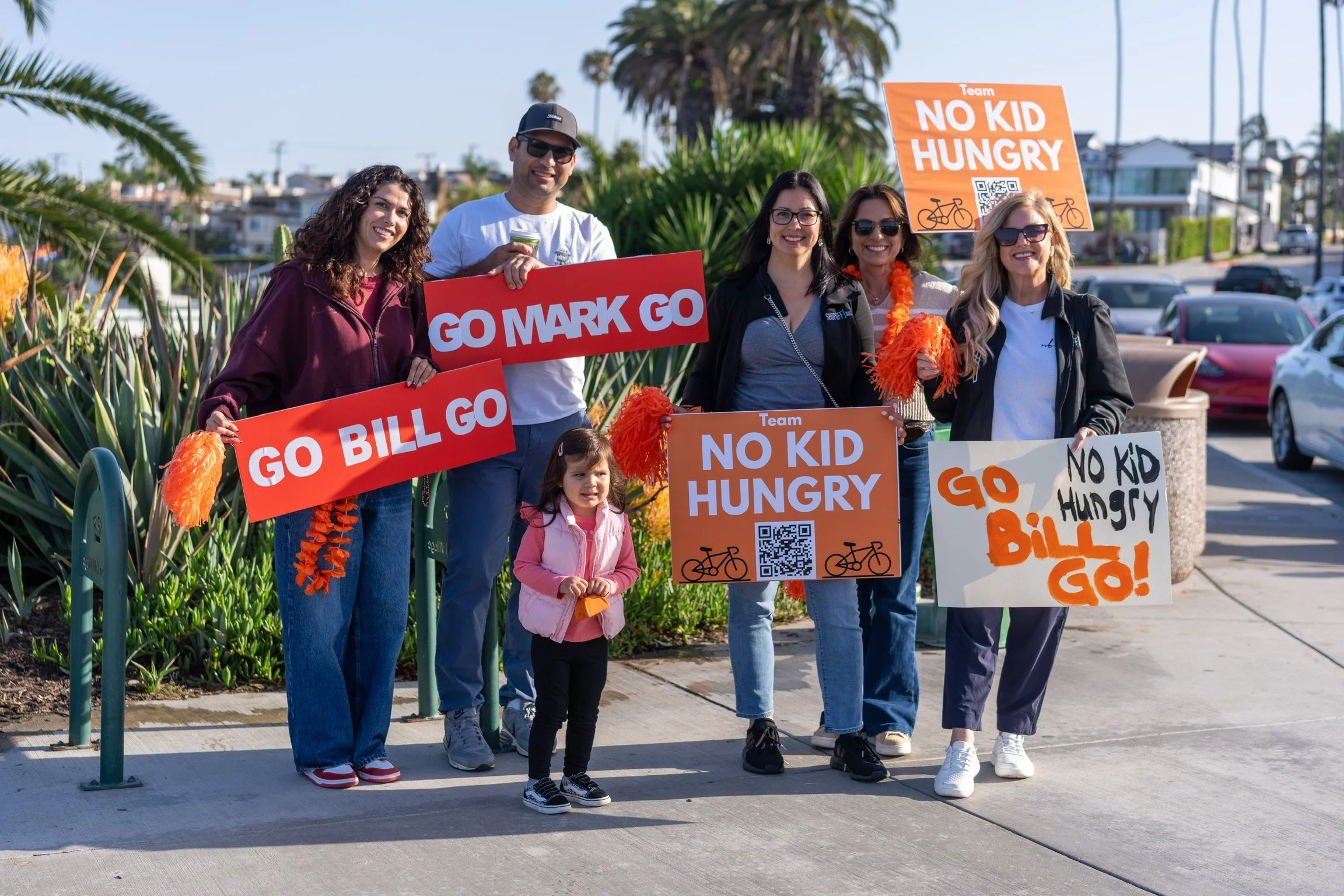 Group of people holding signs at a protest or rally. The signs include messages like 'GO BILL GO', 'GO MARK GO', and 'NO KID HUNGRY'. The setting is outdoors with greenery and parked cars in the background.