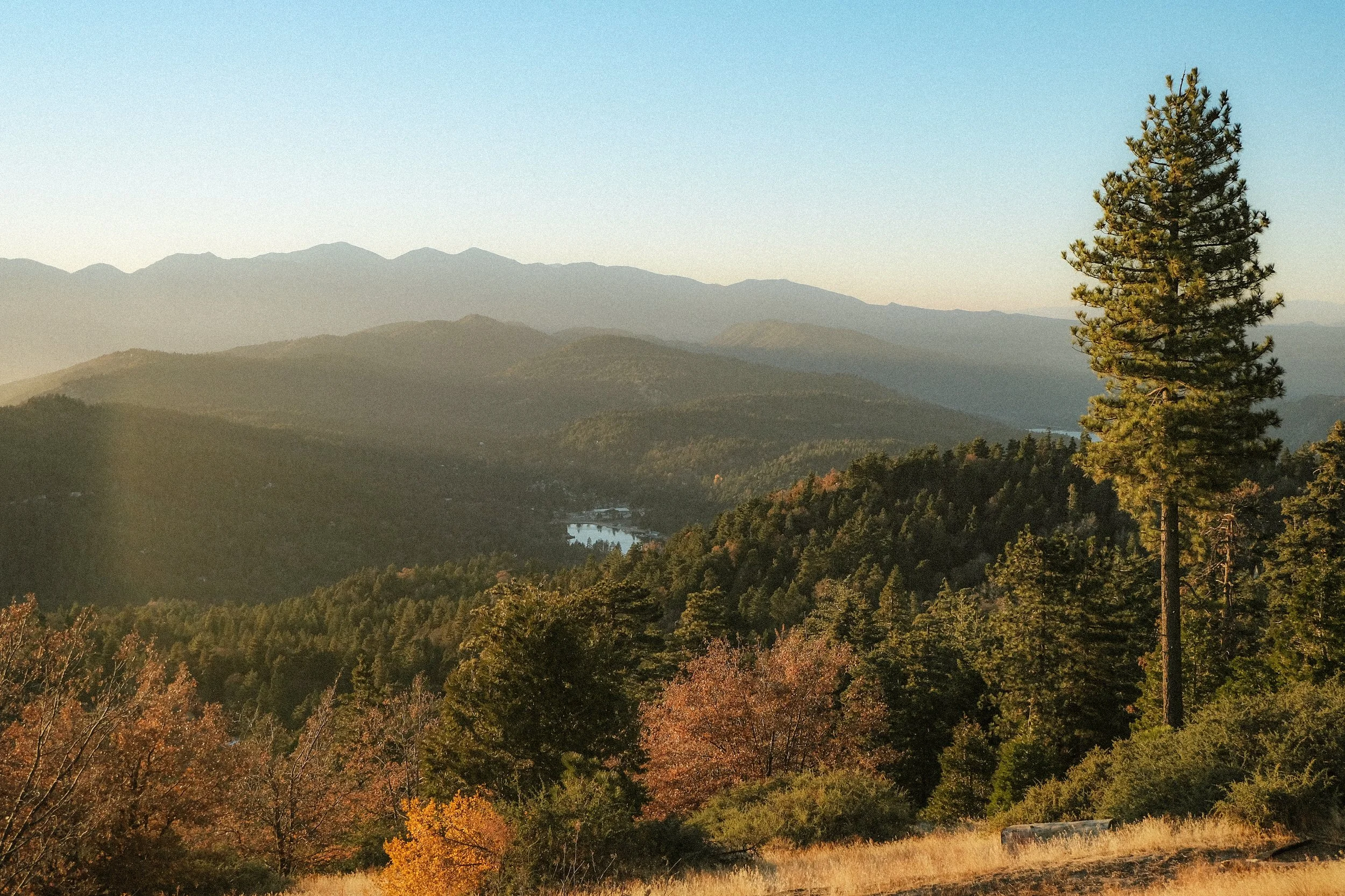 A scenic view of a mountain landscape with trees in fall colors, a lake in the distance, and mountains under a clear sky.