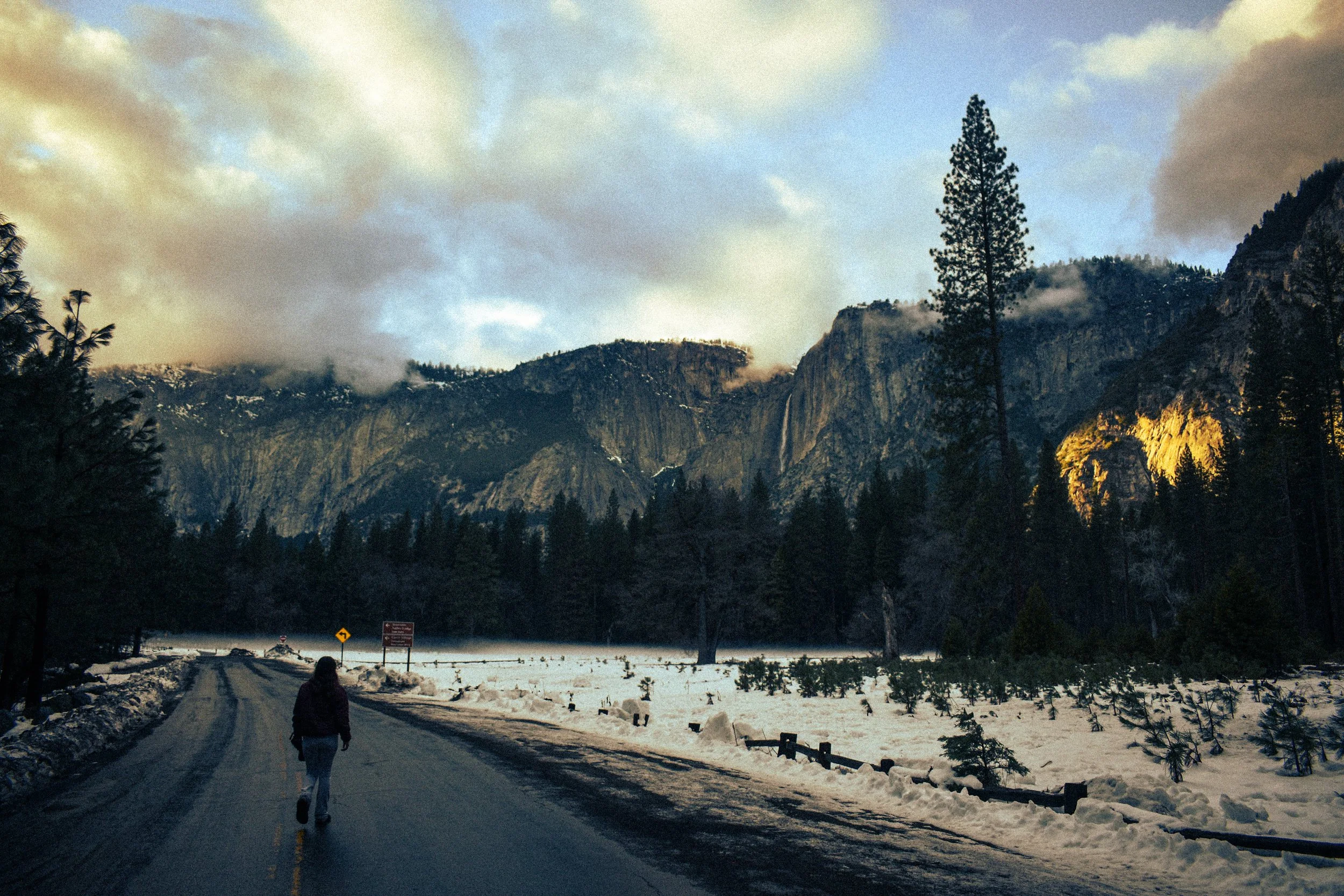 A person walking on a snow-covered road in a mountain landscape, with towering mountains, trees, and a partly cloudy sky in the background.