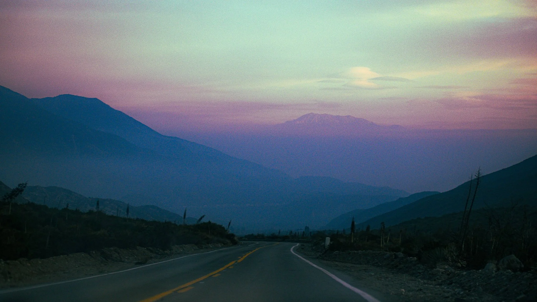 A winding mountain road at dusk with layered mountains in the background, and a pastel-colored sky.