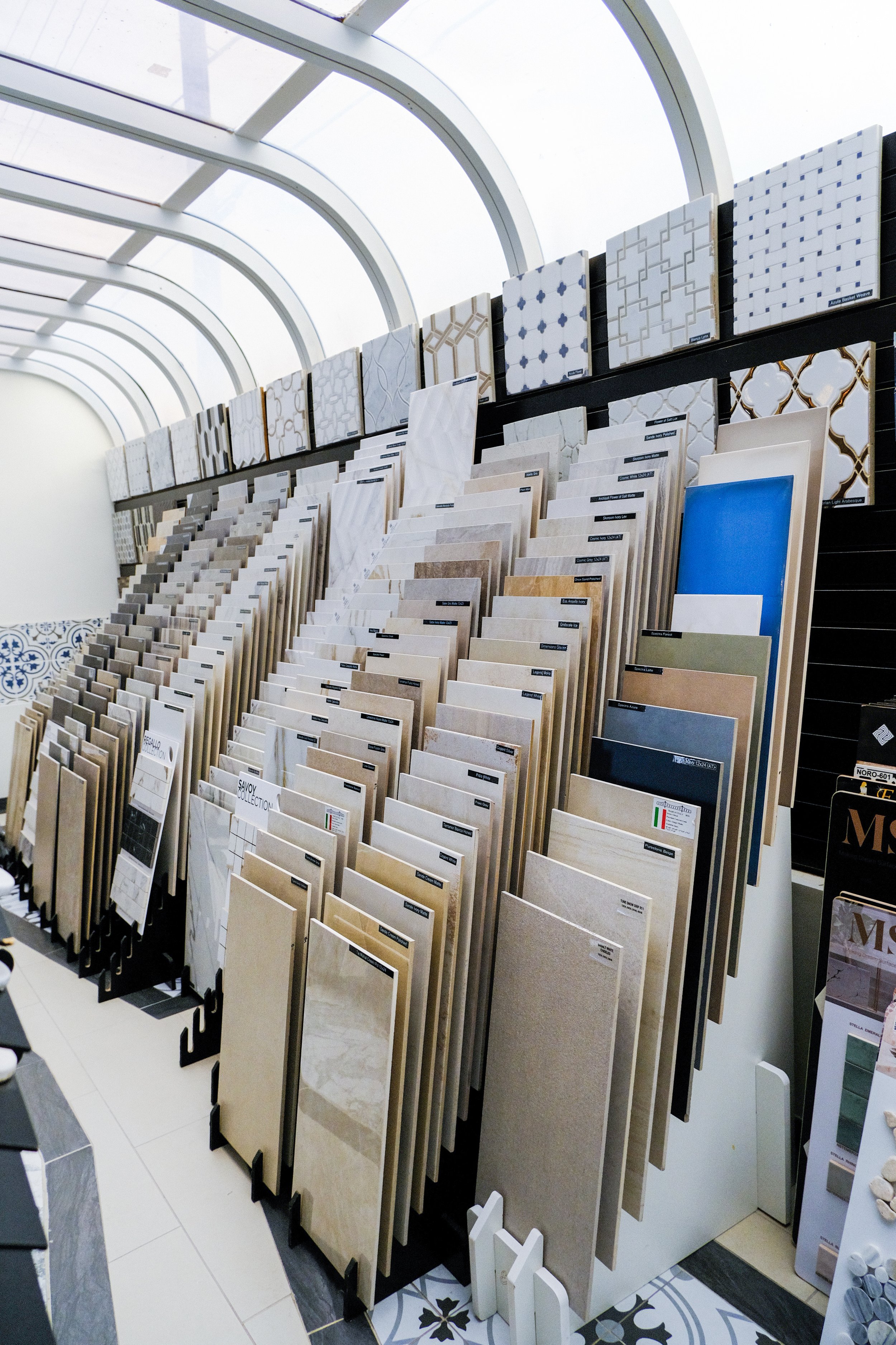 Display of various tiles and flooring samples in a showroom with natural light and a curved glass ceiling.