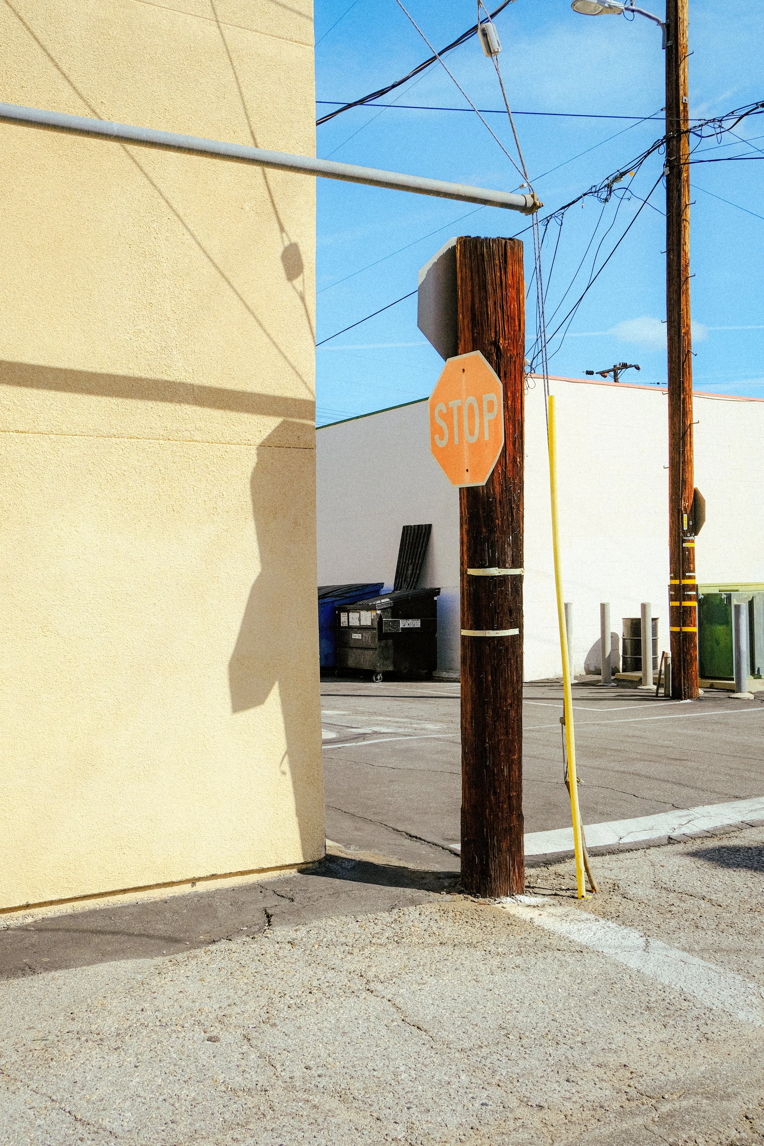 Street corner with a stop sign on a wooden utility pole, yellow wall on the left, parking lot with trash bins behind, utility wires overhead, and a bright blue sky.