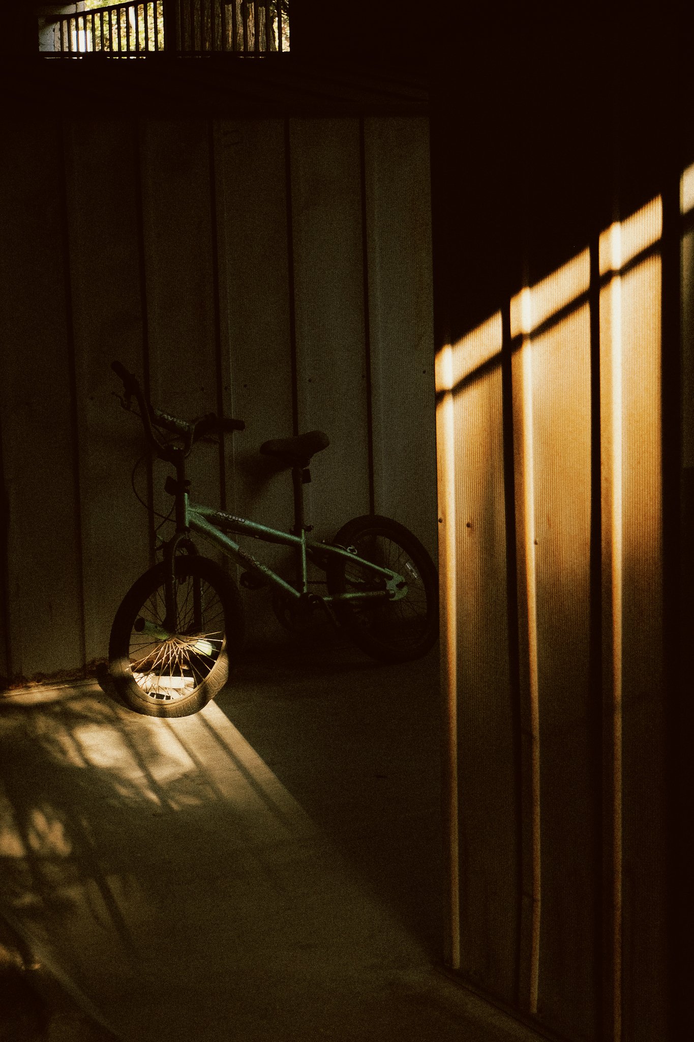 A bicycle leaning against a wall, with sunlight casting shadows through a railing onto the wall and floor inside a dimly lit space.