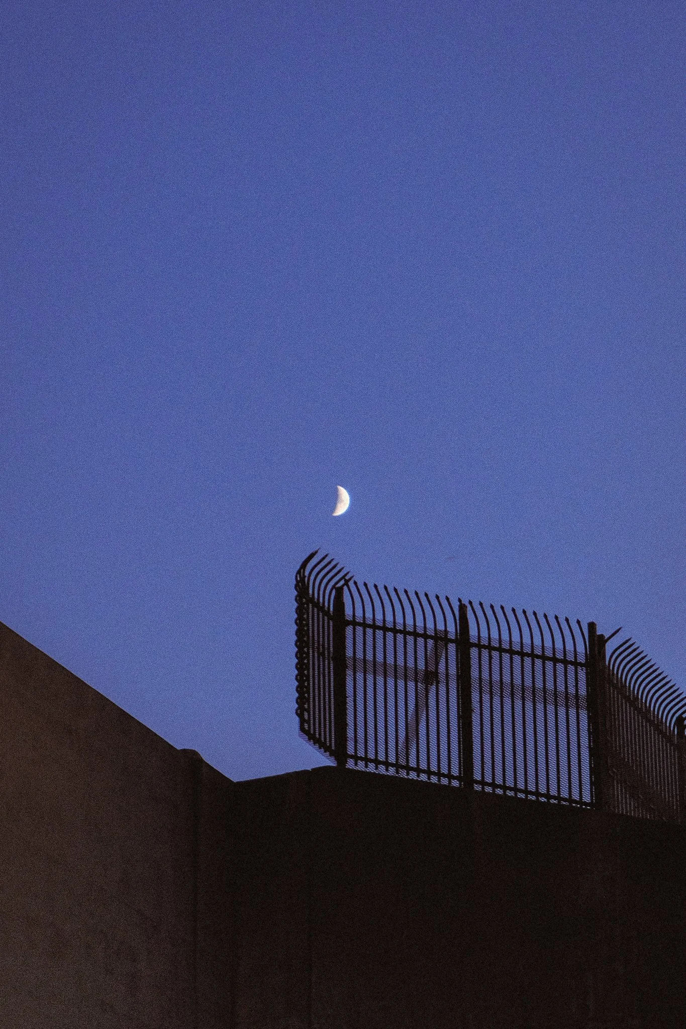 Nighttime sky with a crescent moon visible above a fence on a building rooftop.