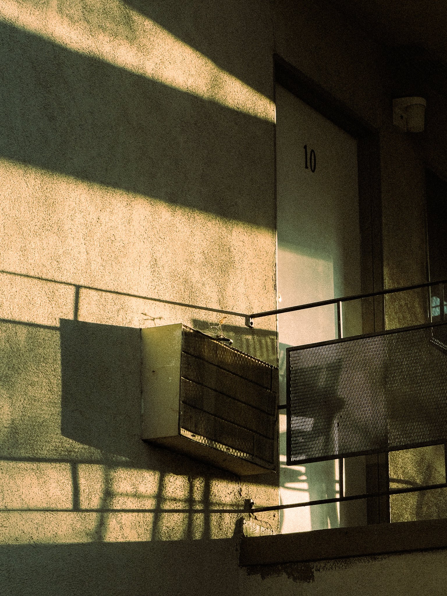 Sunlight casts shadows on a beige wall with a door numbered 10, and a metal grille or vent nearby.