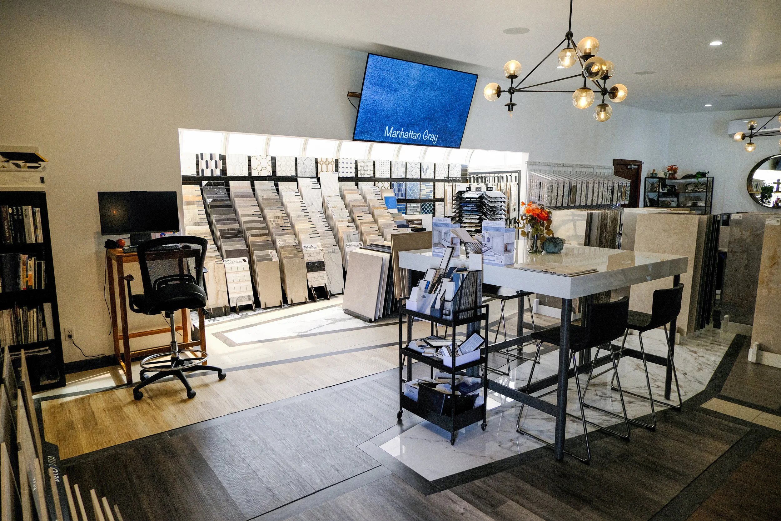 Interior of a tile or flooring store with displays of various tile samples, a small desk with chairs, a magic clock, and modern lighting fixtures.