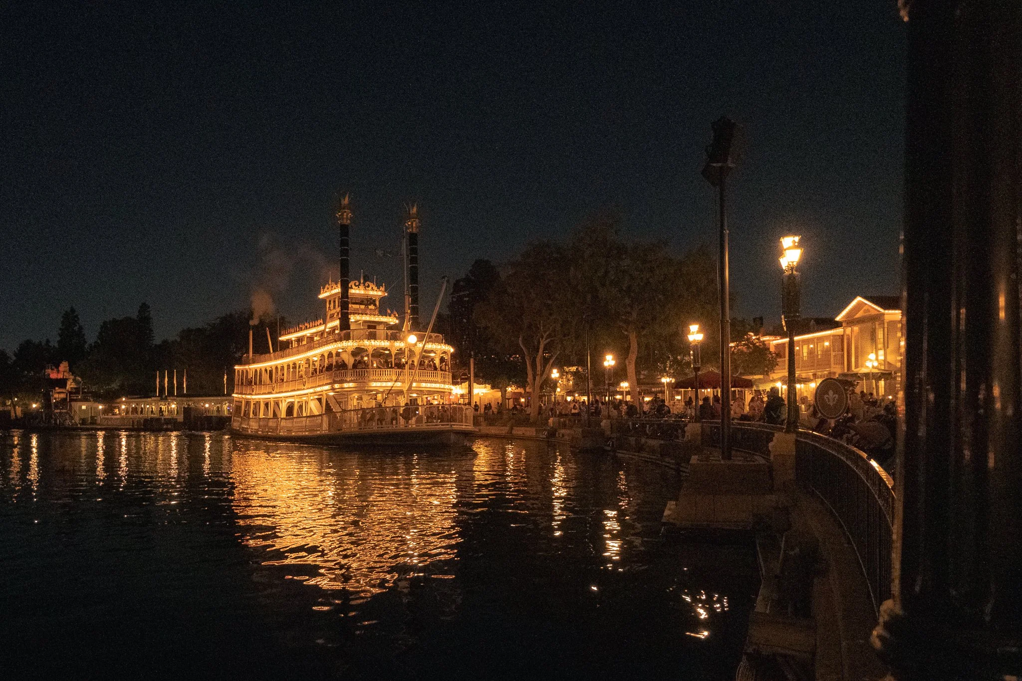 Nighttime view of a lit-up boat docked on a river with a promenade, trees, and buildings illuminated by street lamps along the water.