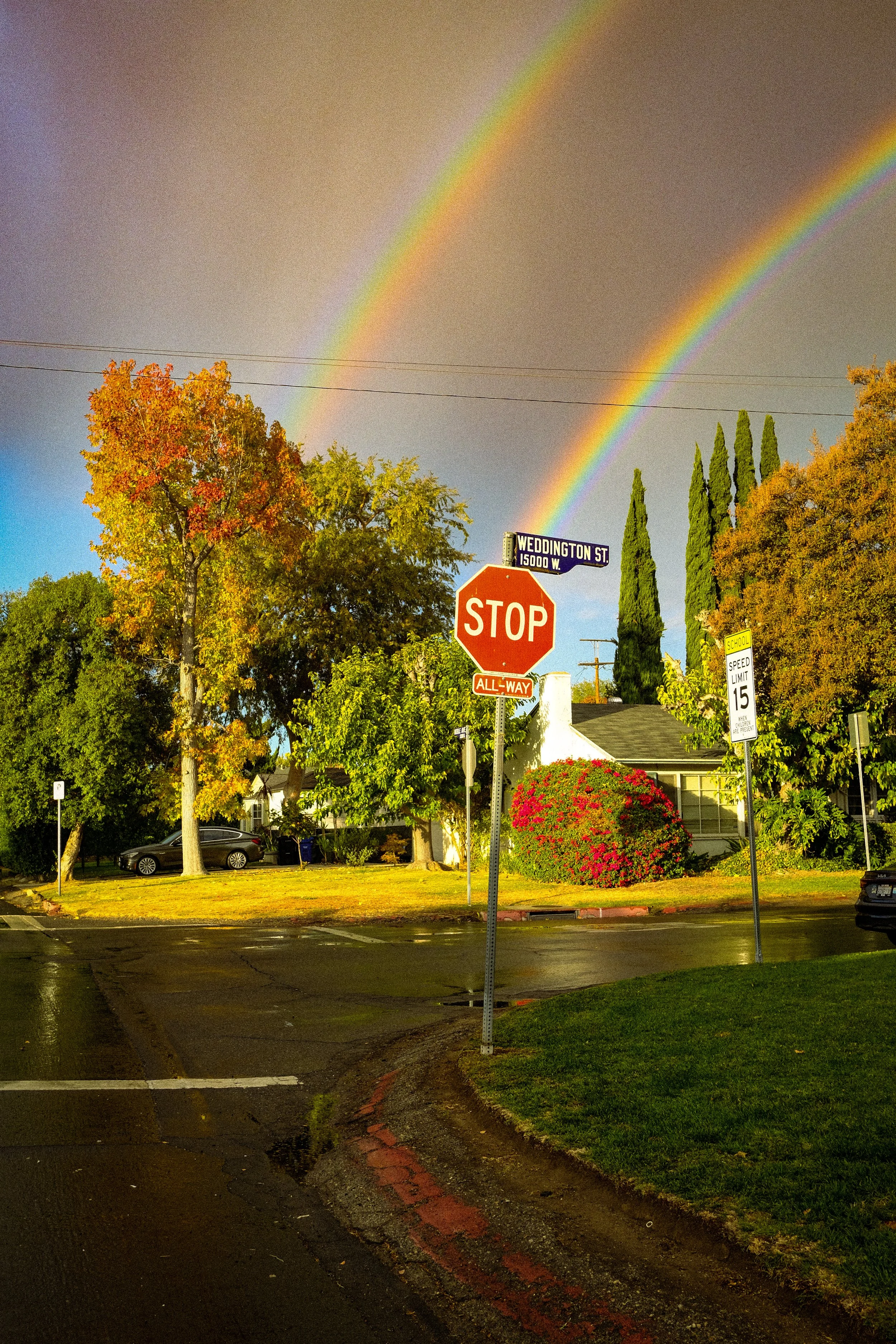 A street intersection with a stop sign, street sign reading 'Weddington St,' and a double rainbow in the sky.