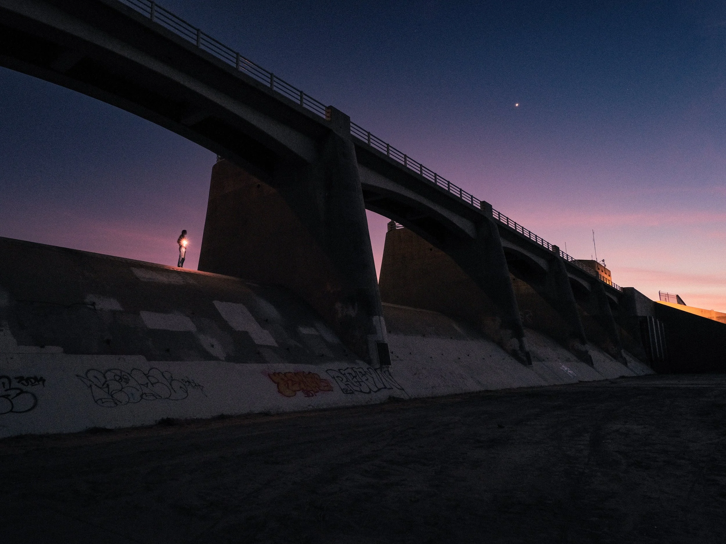 A person standing under a large concrete dam at dusk, holding a flashlight, with a graffiti-covered base and a sky with a few stars visible.